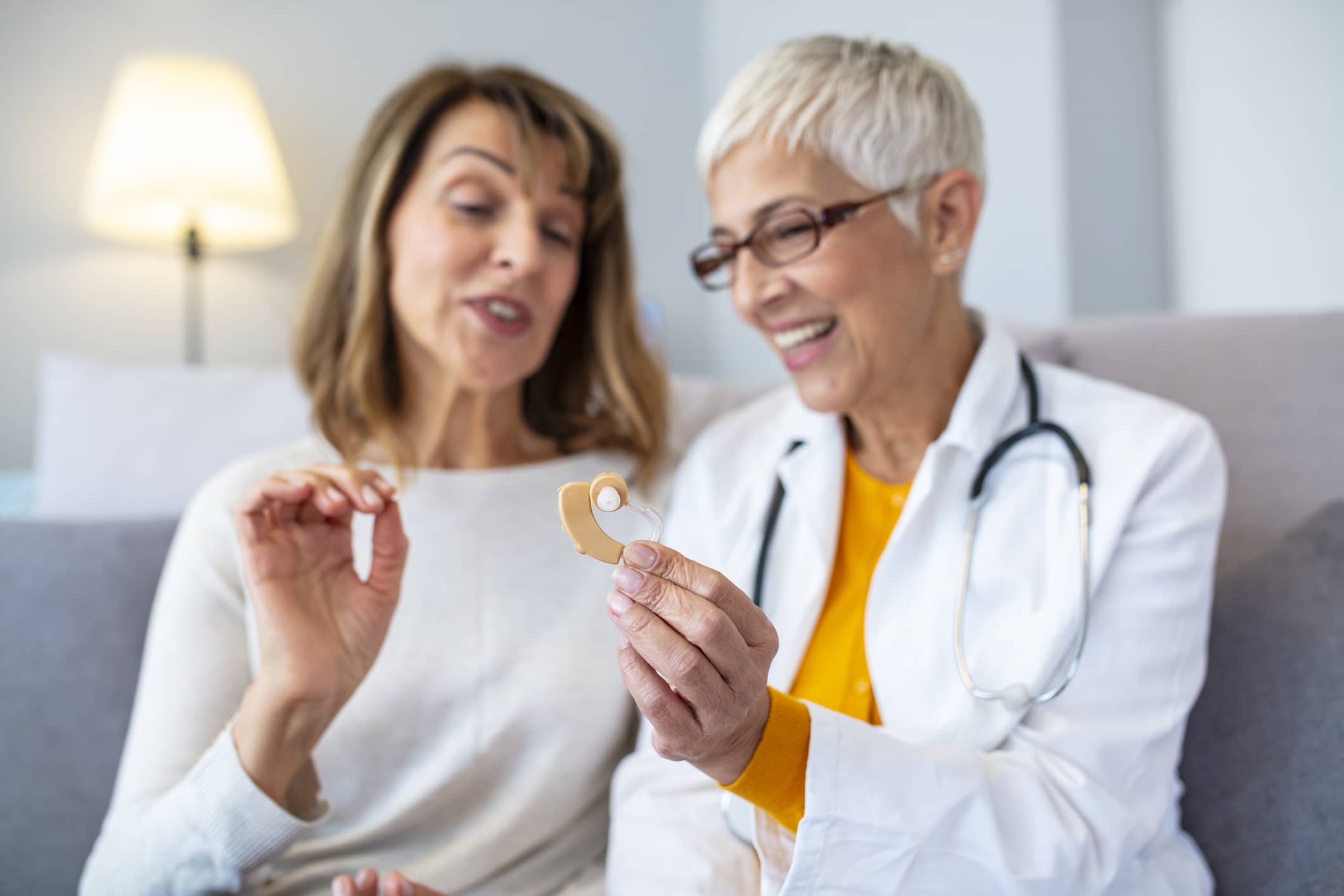 The choice of hearing aid in the doctor's office Mature female doctor hearing specialist in her office trying hearing aid equipment to a patient elderly senior woman. The doctor laryngologist explains to senior woman how to wear a hearing aid