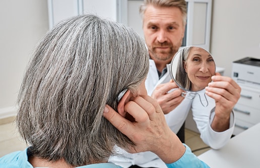 Senior woman trying on a hearing aid while looking into a mirror held by a hearing aid specialist in audiology clinic. Stylish hearing device